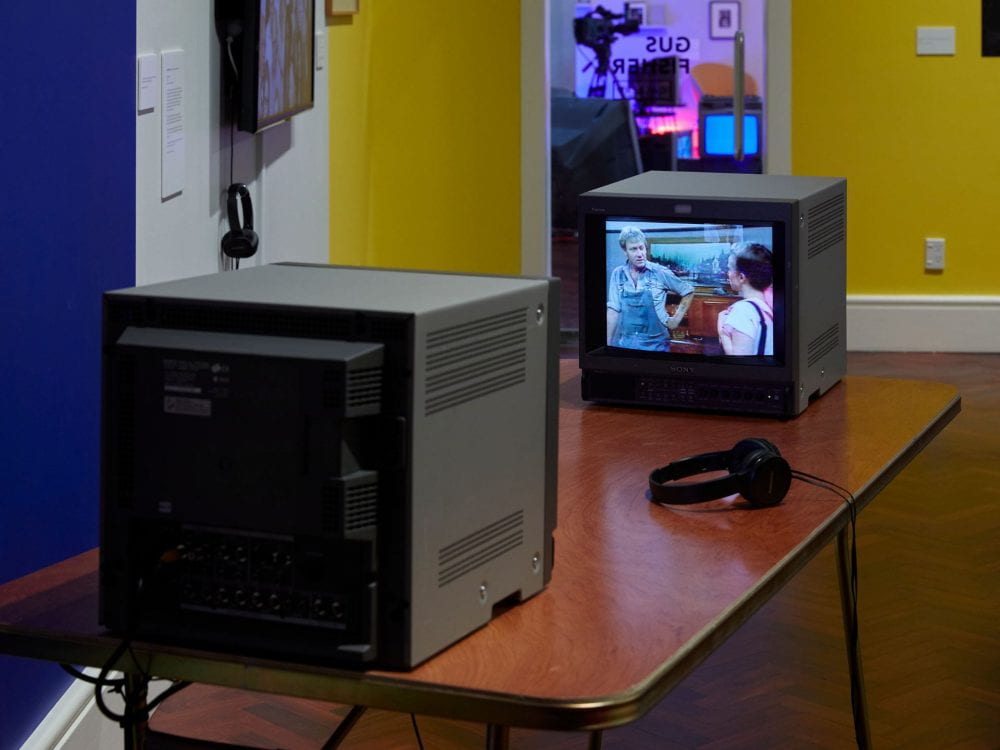 Two CRT television sets face towards each other on a narrow table. On one TV, 1980s clips from soap opera Coronation Street play. On the other TV, the artist sits dressed as an interviewer.