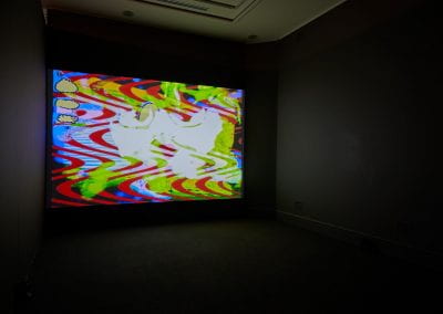 A dark room with a colourful film projected onto one wall. A man's face is visible in the film, fading into psychedelic wave patterns.