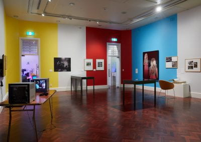 A gallery room with yellow, white, red and blue walls. Various artworks are displayed across the space.