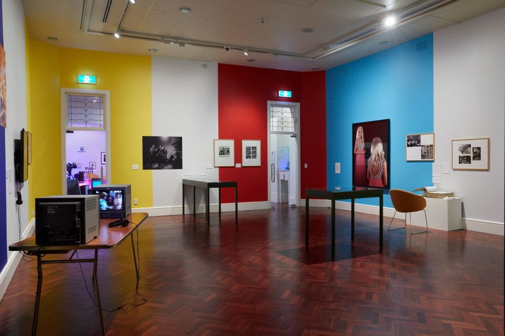 A gallery room with yellow, white, red and blue walls. Various artworks are displayed across the space.
