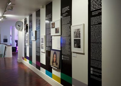 A series of black and white vinyls running vertically down a corridor wall, with framed photographs and archived objects displayed alongside.