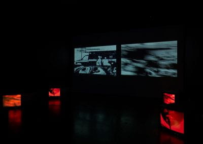 Two films projected side by side in a dark room, with four CRT television sets positioned in front on the floor. On the CRT TVs a person eating noodles is depicted, entirely in red.
