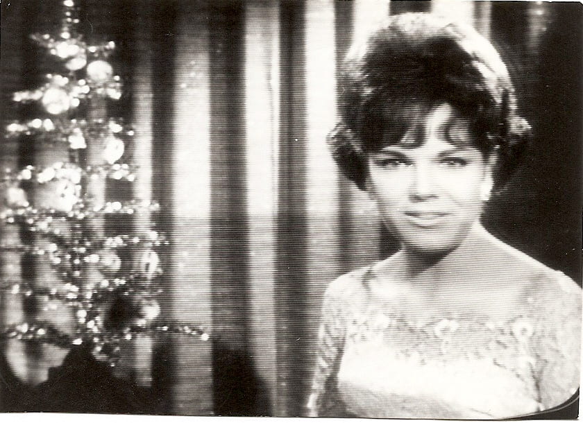 A young Jennie Goodwin, dressed in a light dress with short dark hair, faces towards the camera presenting. Next to her is a Christmas tree.