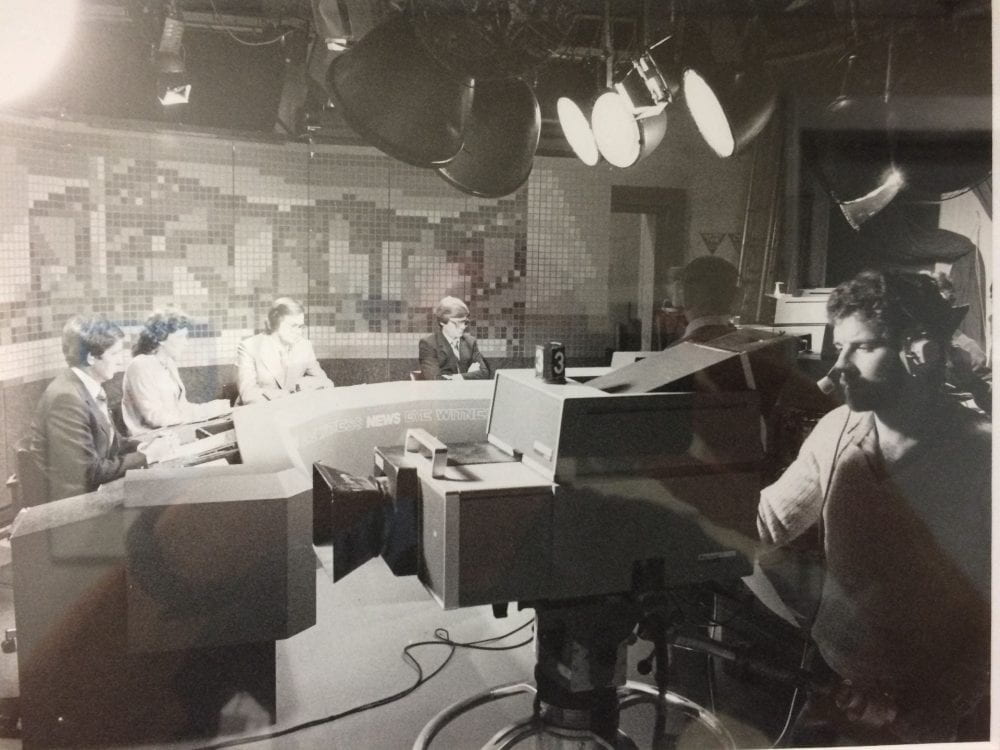 Interior of a television studio. A cameraman points a large camera on a stand to the left. Just beyond him, two presenters sit at a news desk preparing for their cue to go live.