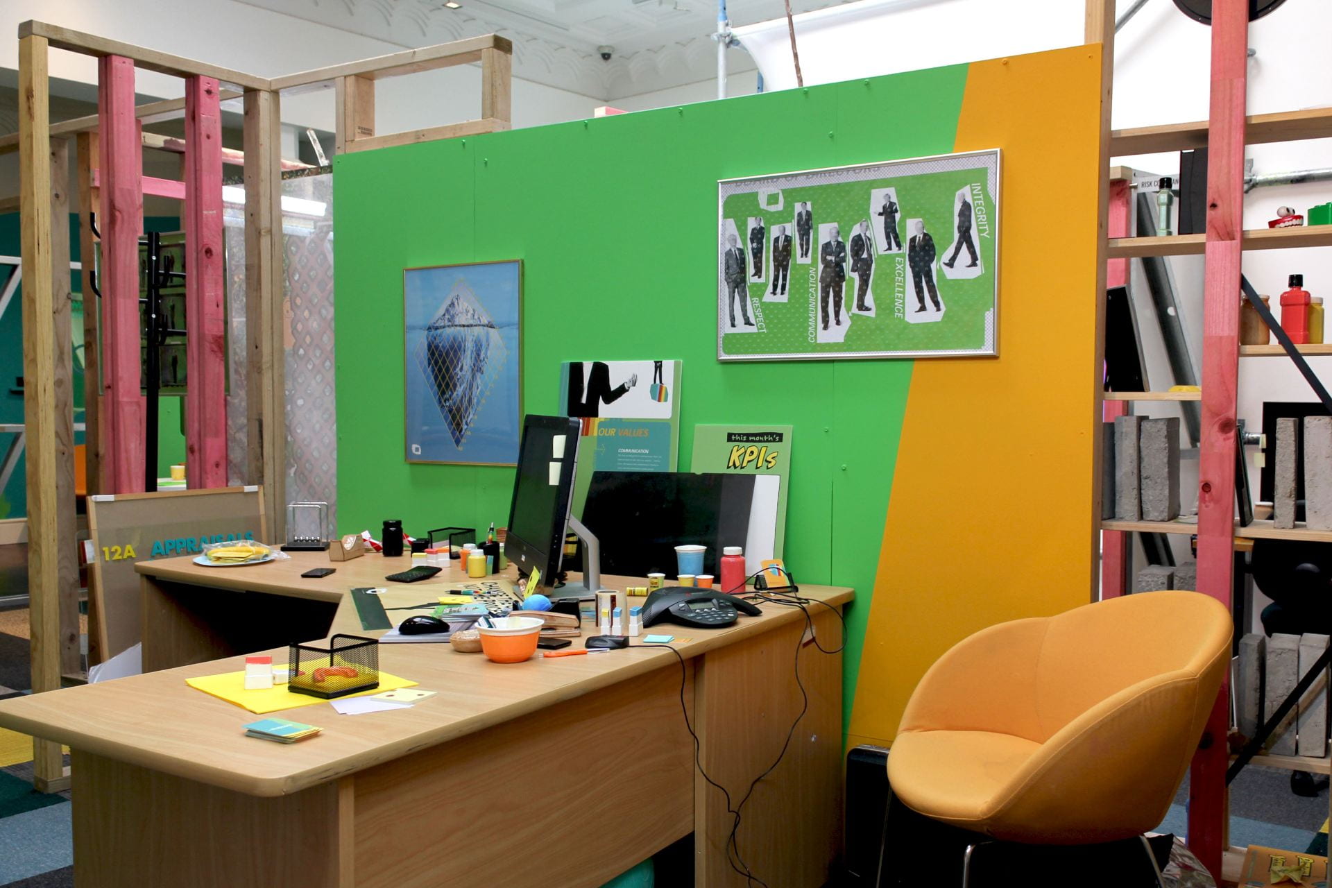 A cluttered office space. A large wooden corner desk sits next to a green and orange wall and an orange chair. It is covered with cast ceramic objects and post-it notes.