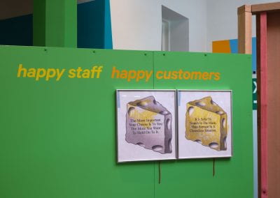 A green wall with text reading "happy staff, happy customers" across it. Two framed photographs of blocks of cheese hang next to each other with text placed in the centre of each. The left hand one reads "the more important the cheese is to you the more you want to hold on to it". The right one reads "it is safer to search in the maze, than to remain in a cheeseless situation".
