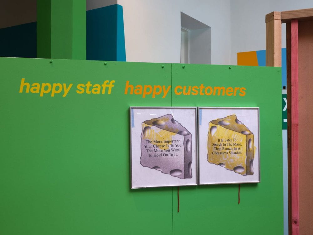 A green wall with text reading "happy staff, happy customers" across it. Two framed photographs of blocks of cheese hang next to each other with text placed in the centre of each. The left hand one reads "the more important the cheese is to you the more you want to hold on to it". The right one reads "it is safer to search in the maze, than to remain in a cheeseless situation".
