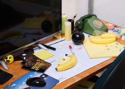 A cluttered office desk. All of the items on the desk - a telephone, a wireless mouse, a banana, a magic 8 ball and pencil cups - have been made out of glazed ceramic.