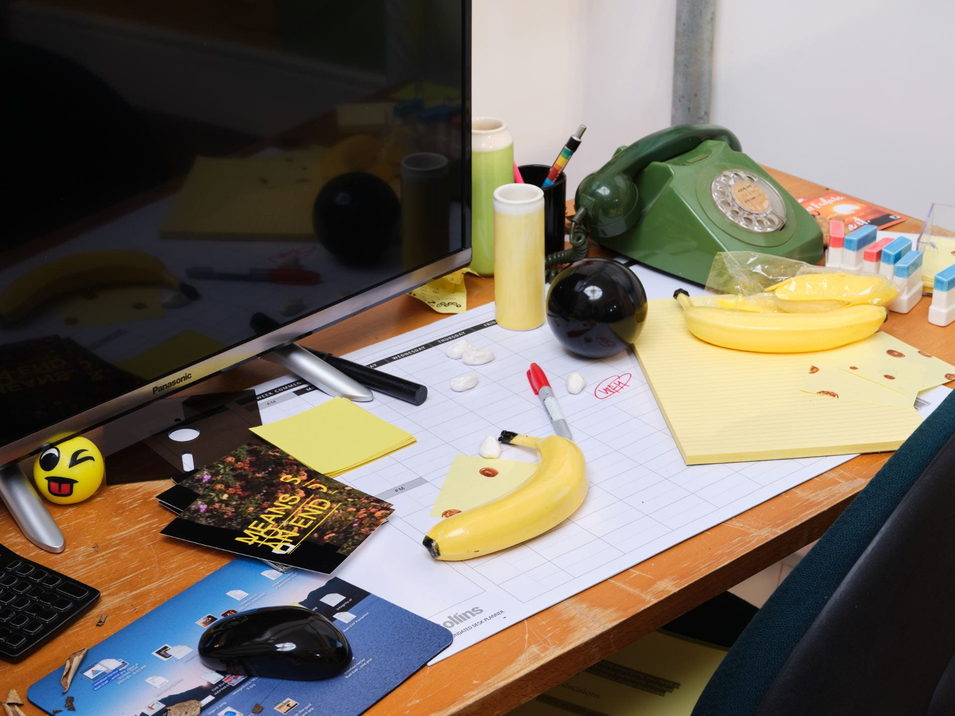 A cluttered office desk. All of the items on the desk - a telephone, a wireless mouse, a banana, a magic 8 ball and pencil cups - have been made out of glazed ceramic.