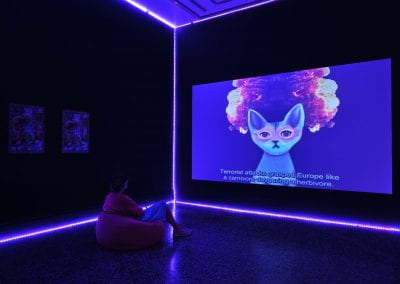 A film projected in a dark room lined with purple neon lights. A woman sitting on a bean bag watches an animated blue and pink cat talk at the viewer, surrounded by an explosion in the background. The subtitle along the bottom of the screen reads "terrorist attacks grasped Europe like a carnivore devouring a herbivore".