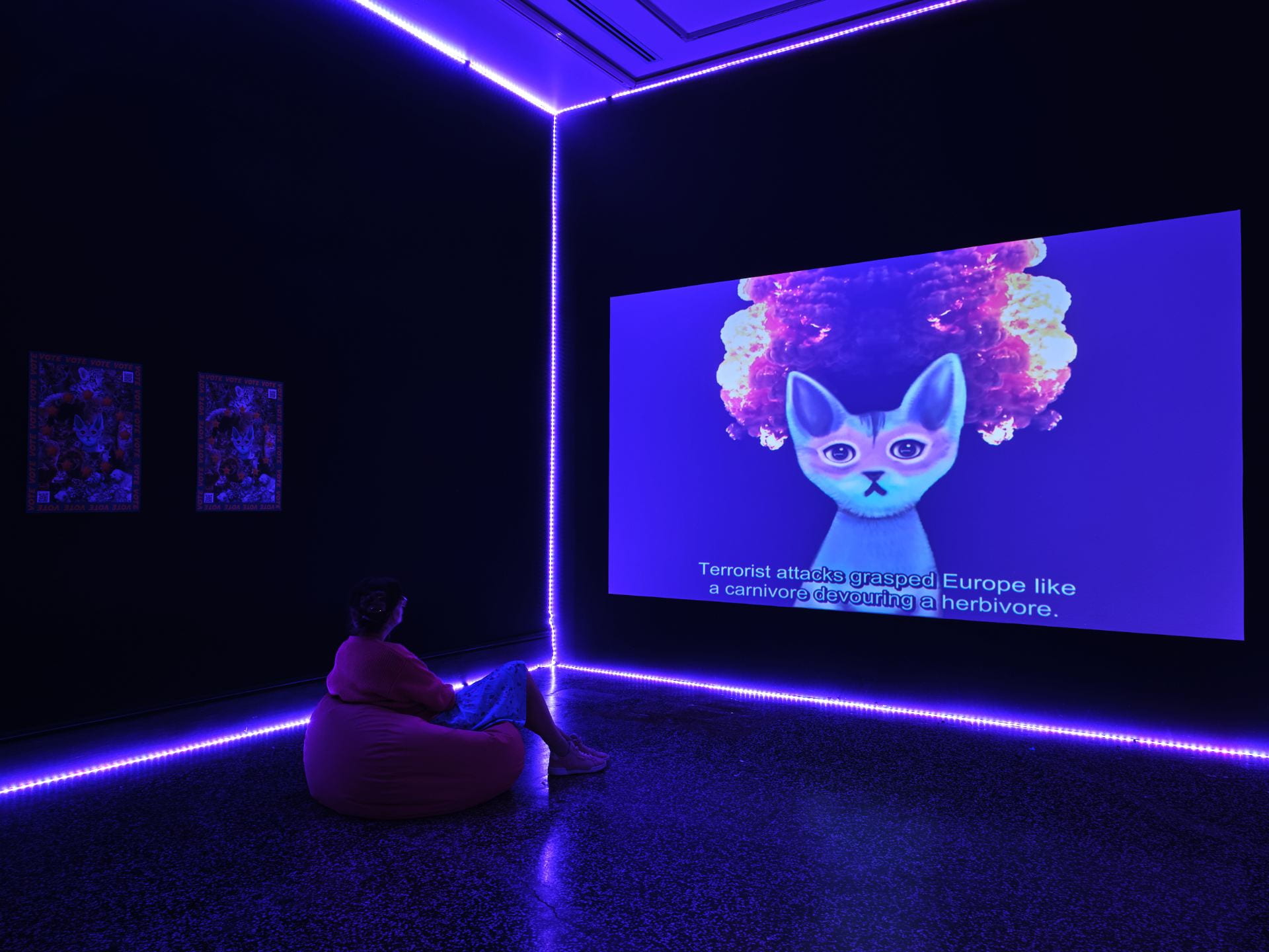 A film projected in a dark room lined with purple neon lights. A woman sitting on a bean bag watches an animated blue and pink cat talk at the viewer, surrounded by an explosion in the background. The subtitle along the bottom of the screen reads "terrorist attacks grasped Europe like a carnivore devouring a herbivore".