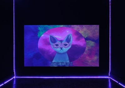 A film projected in a dark room lined with purple neon lights. An animated blue and pink cat talks at the camera, surrounded by a galaxy landscape. The subtitle along the bottom of the screen reads "curiosity kills the cat, and trains the Kitty AI".