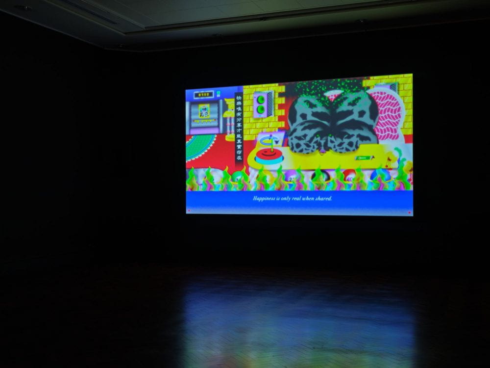 A film projected in a dark room. The film shows a bright animation of a cartoon cow surrounded by green flames, slow cooked into high-quality beef. The subtitle along the bottom of the film reads "happiness is only real when shared".