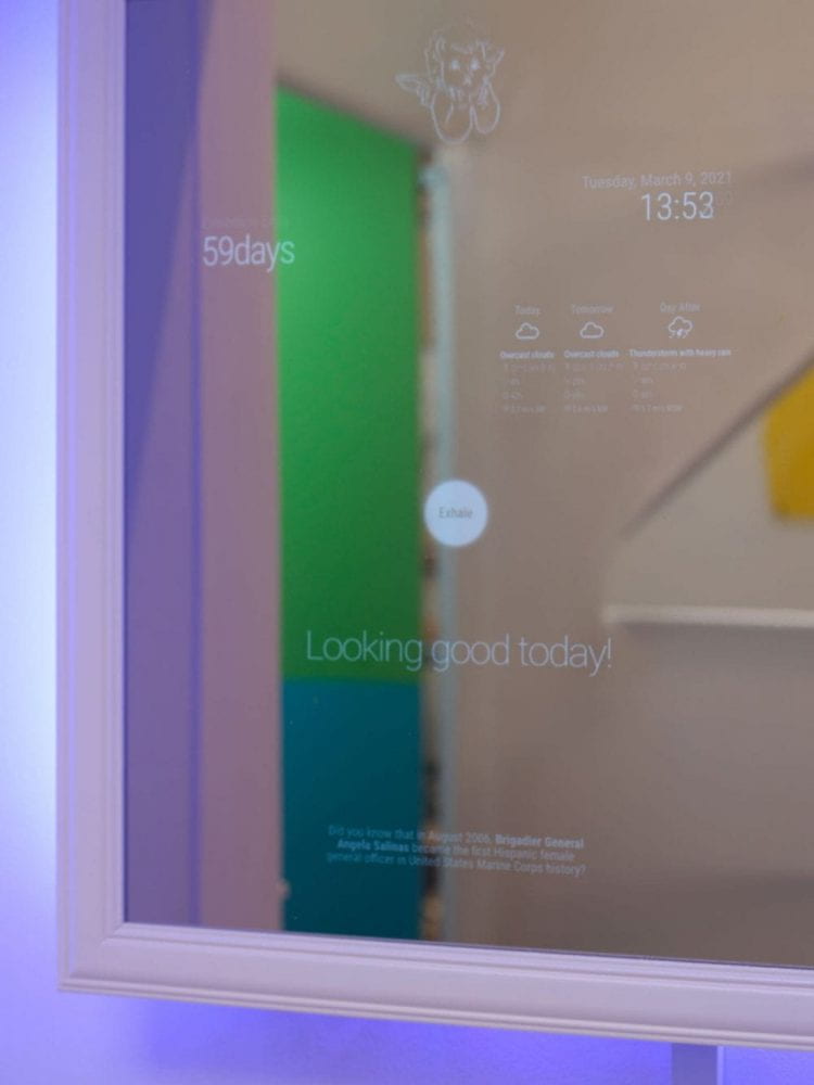 A framed smart mirror with weather reports, self-care reminders and compliments overlaid on its face, on a wall surrounded by purple lighting