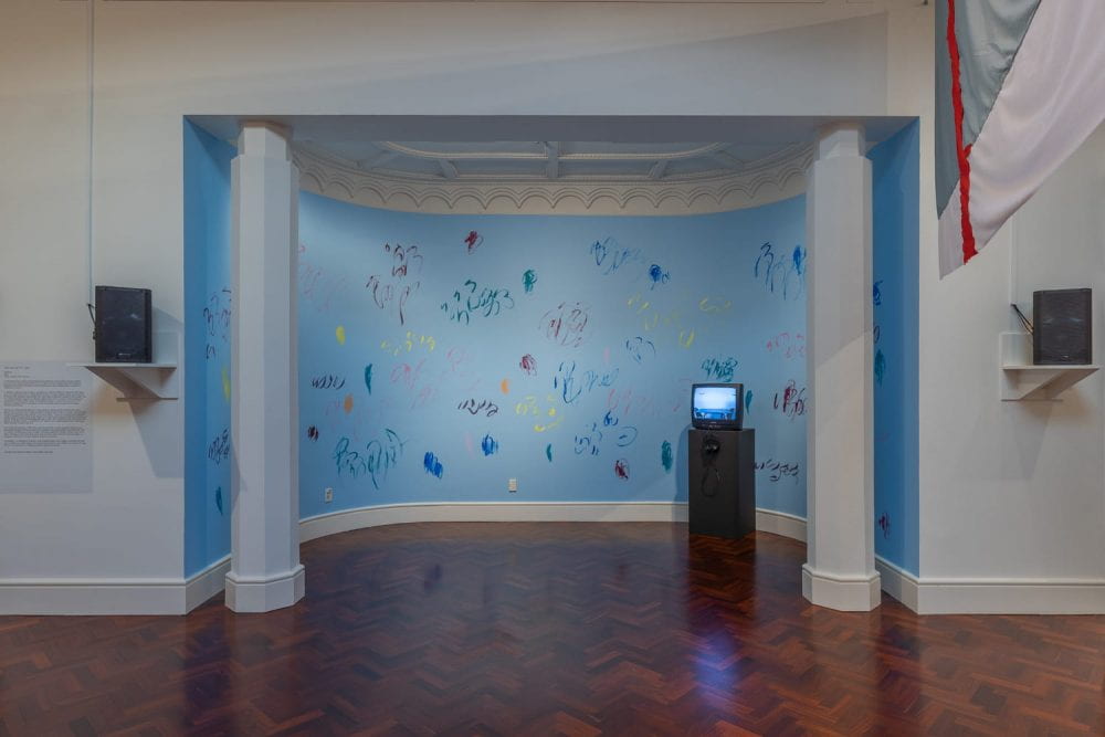 An alcove space painted blue, with colourful mark-making and squiggles painted all over it. A CRT television sits on a black plinth in the alcove. Speakers are installed on shelves flanking the alcove.