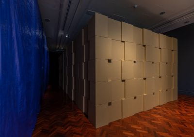 A dimly lit room filled with a large rectangular structure, made out of stacked cardboard boxes. A blue tarpaulin hangs on the left wall.