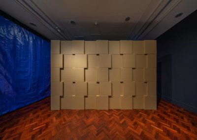 Cardboard boxes are stacked on top of each other to form a large rectangular structure, in a dark gallery room. Some of the boxes are pushed out slightly to create a pattern.