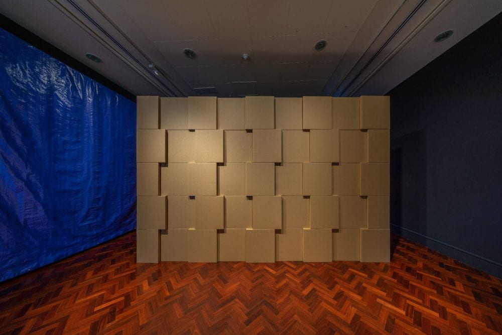 Cardboard boxes are stacked on top of each other to form a large rectangular structure, in a dark gallery room. Some of the boxes are pushed out slightly to create a pattern.