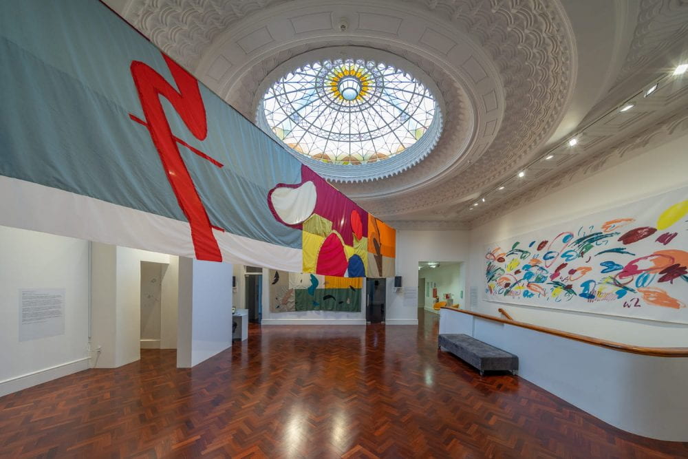 A large gallery space with an elaborate stained glass dome ceiling, and herringbone wooden floor. A colourful fabric banner stretches across the upper half of the room. On the right-hand wall, a long painting of colourful abstract shapes and squiggles hangs over a stairwell.