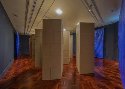 A room filled with vertical stacks of cardboard boxes, arranged into tall towers. Three blue tarpaulins hang on black walls around the room.