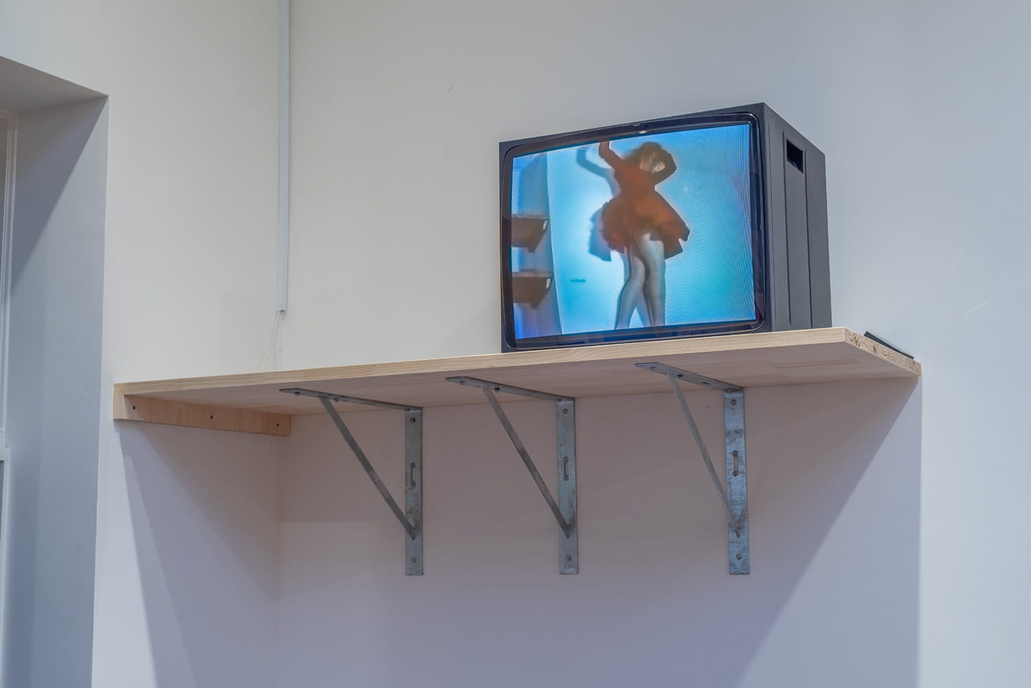 A wooden shelf with a large CRT television set on top of it. On the TV, a film plays showing a woman in a red dress climbing around a small apartment.