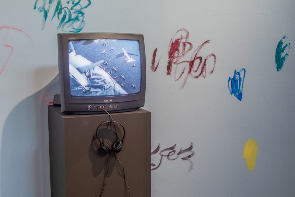 An old CRT television set sits on top of a black plinth. On the TV a film plays showing miniature aeroplanes flying above a bed. The wall behind the TV is blue with abstract colourful squiggles painted onto it.