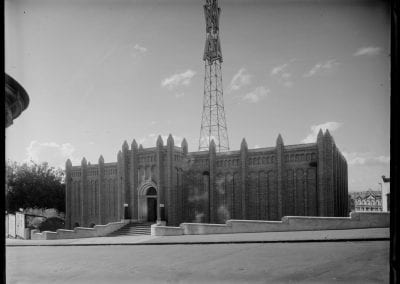 An old black and white photograph of an elaborate brick building, taken from the street, with a tall radio mast which has neon letters '1YA' displayed on it.
