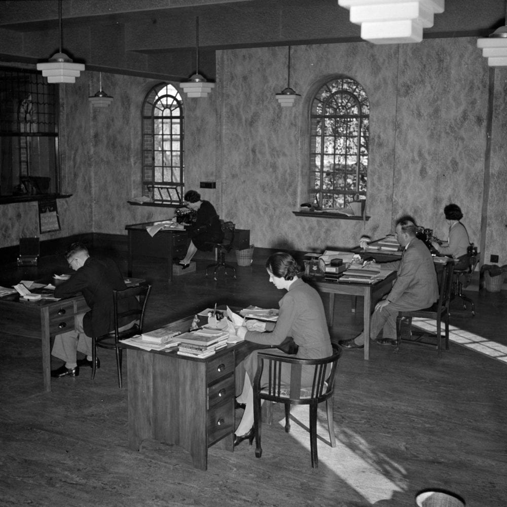 Office workers sit at desks in an open-plan room, fitted in Art Deco interior design.