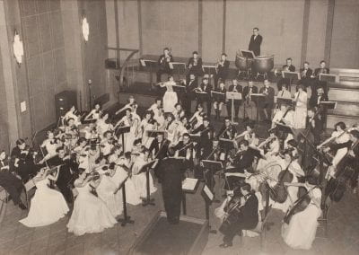 An orchestra dressed in formal attire plays in a large studio room.