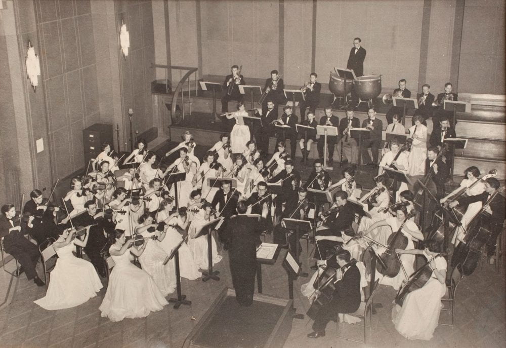 An orchestra dressed in formal attire plays in a large studio room.