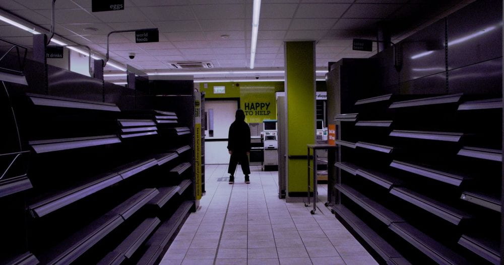 A figure fully clad in black stands in a dimly lit, abandoned co-op amongst empty shelves.
