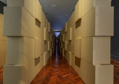 Looking down a v-shaped path created using stacked cardboard boxes, inside a gallery space.