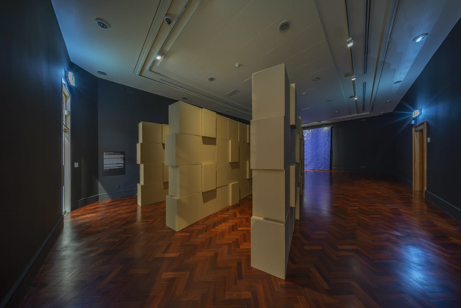 A room with three rows of stacked cardboard boxes arranged in a v-shape, pointed towards a blue tarpaulin hanging on the back wall.