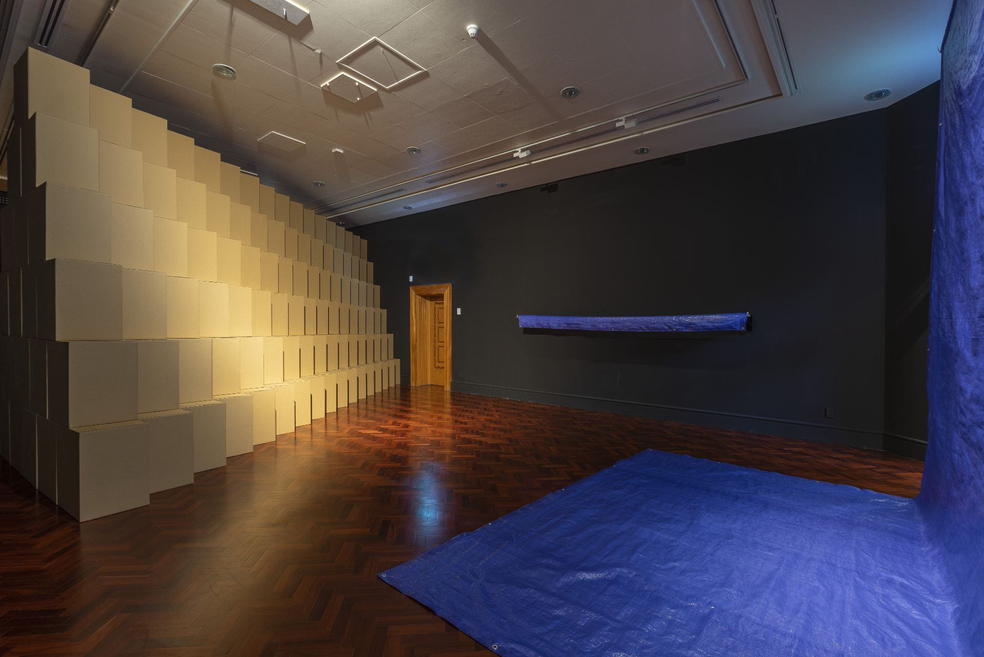 A dramatically lit gallery filled with stacked cardboard boxes in a tiered pattern. A blue tarpaulin is rolled up on one black wall, and another spreads out onto the wooden floor.