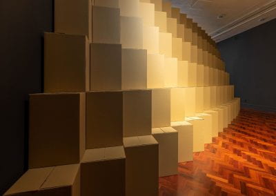 A dramatically lit gallery filled with stacked cardboard boxes in a tiered pattern.