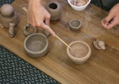 A hand holding a stick presses ridges into the rim of a clay pot, with other clay pots surrounding them on a wooden board.