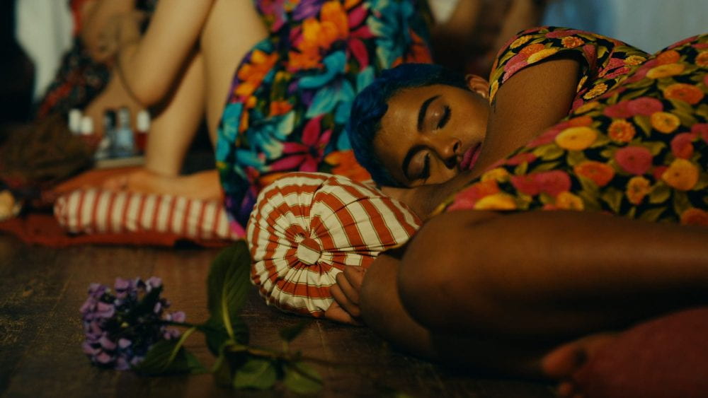 A person in a floral patterned dress sleeps on a striped cushion, next to another person in a patterned dress.
