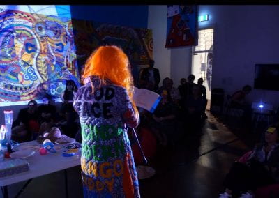 A person in a bright orange wig and quilted coat performs to a crowd in a dark room, lit by projected patterns.