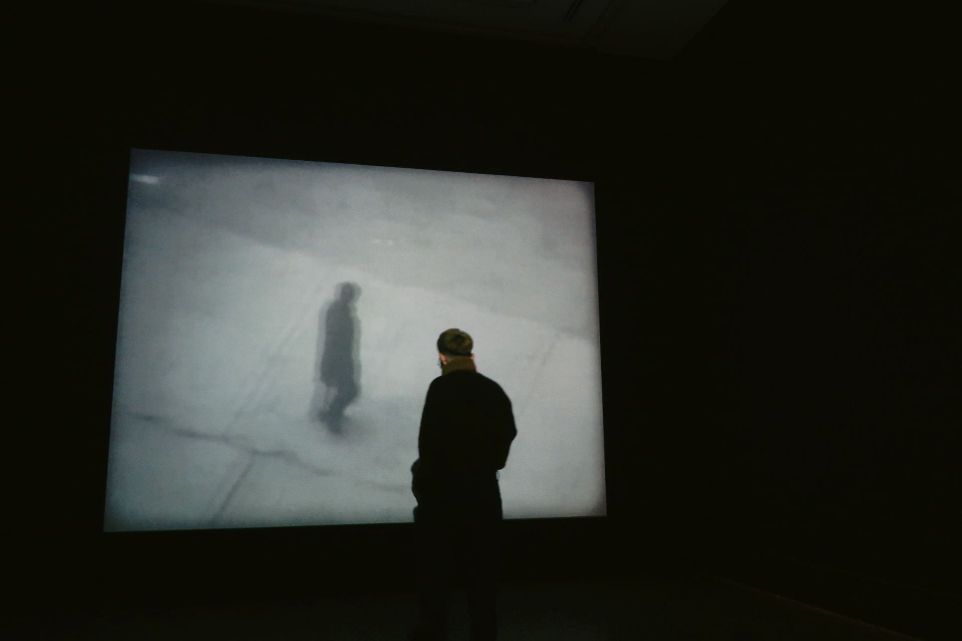 A person in a black coat, standing in a dark room, watches a black and white film.