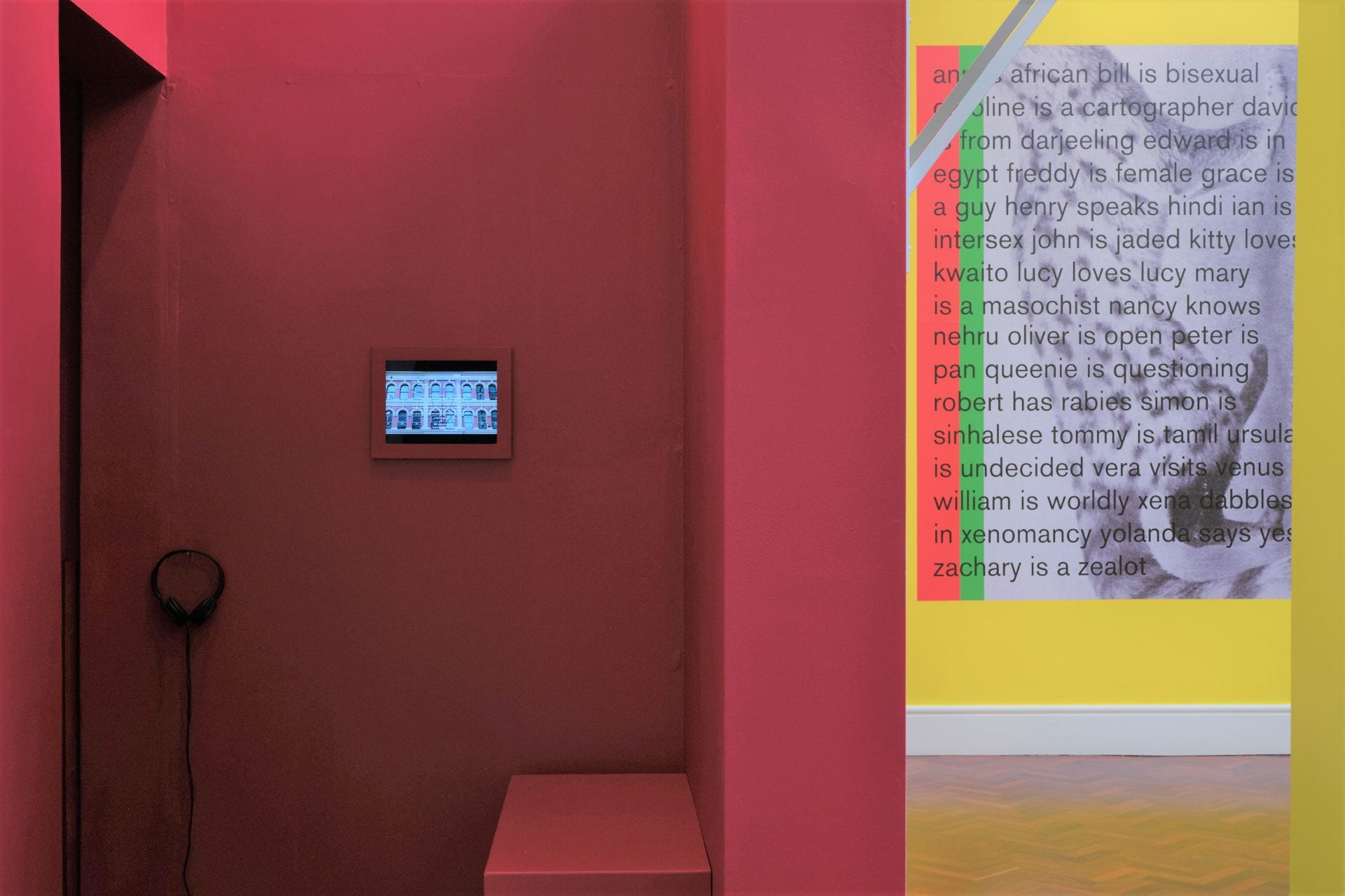 A red booth space with a small iPad playing a film mounted on the wall. In the background is a large grey, red and green wallpaper on a yellow wall.
