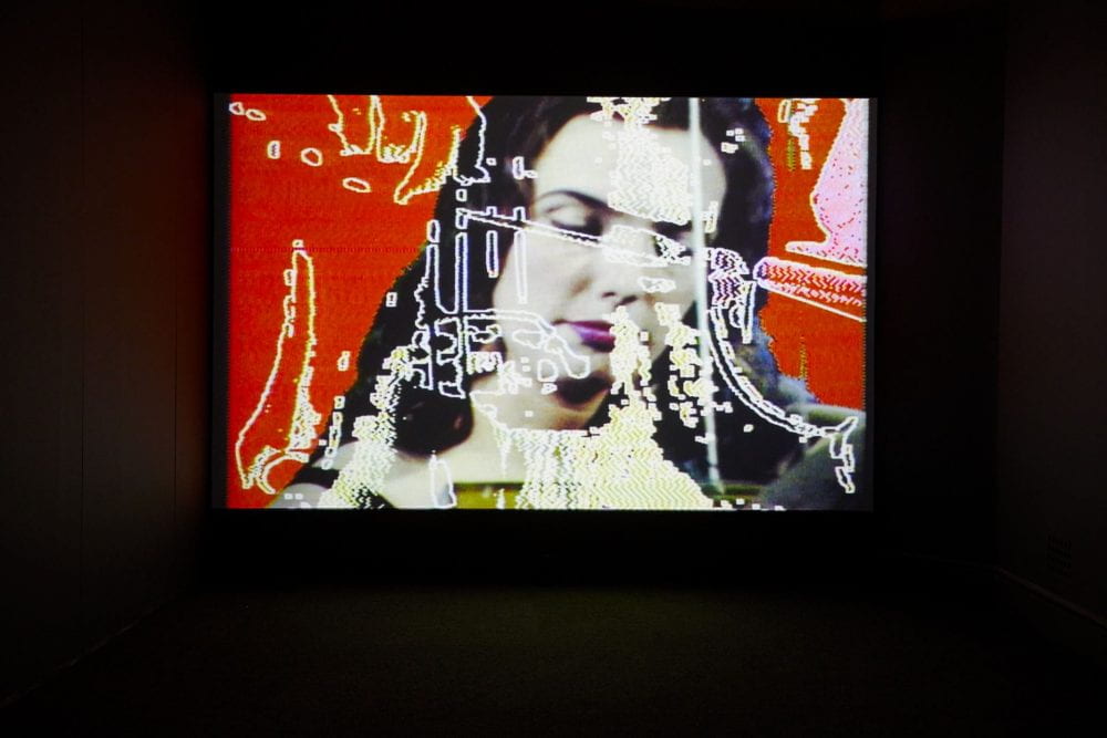 A film projected in a dark room. The film shows a woman's face against a red backdrop, overlaid with abstract patterns.