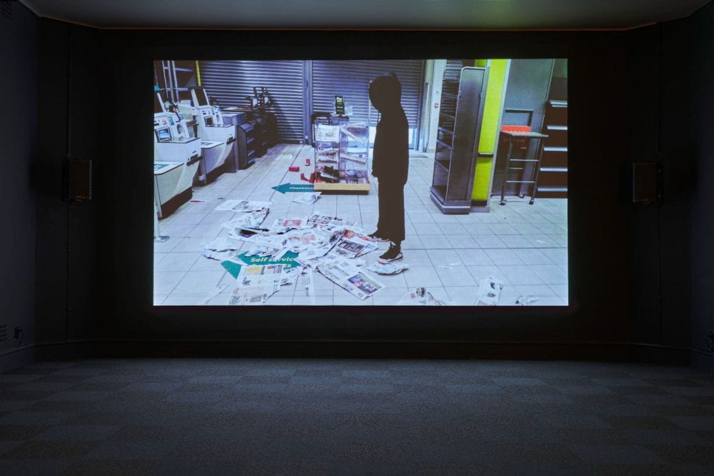 A dark room showing a film projected on the back wall. In the film, a figure clothed in black dances through a deserted co-op store.