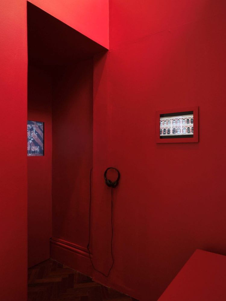 A bright red, tall narrow booth space. On the right-hand wall is a small screen playing a film which depicts heritage buildings in Auckland's CBD. To its left is a pair of headphones. Further left, a larger film can faintly be seen.