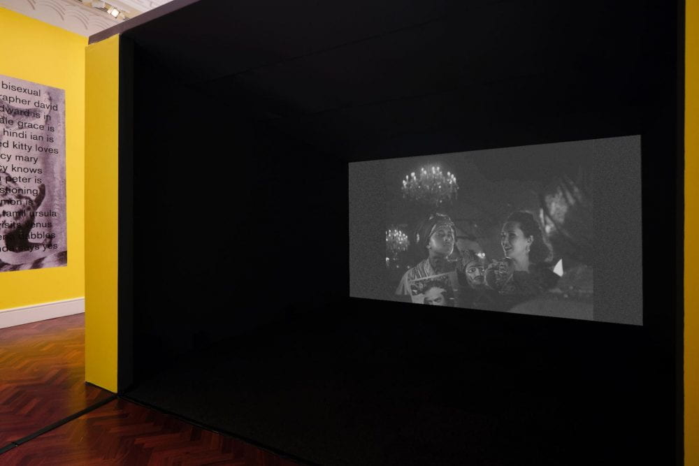 A large enclosed film booth painted yellow and black. The film projected inside depicts a nonbinary person and a woman seated at a dressing room table, surrounded by elaborate costumes, props and a glowing chandelier. The film is in black and white.