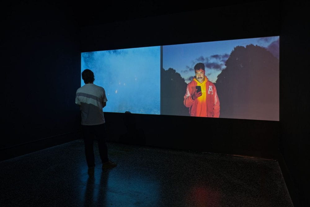A two-channel film projected in a dark room. A man stands on the left hand side watching the films. The left film depicts a smoky atmosphere, and the right film shows a man dressed in orange staring at his phone in a dusky outdoor setting.