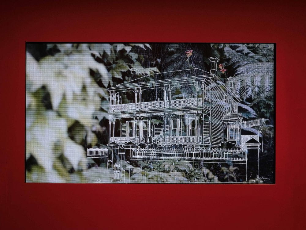 A film screen embedded into a bright red wall. The film shows a line drawing of a multi-storeyed villa, against a background of foliage.