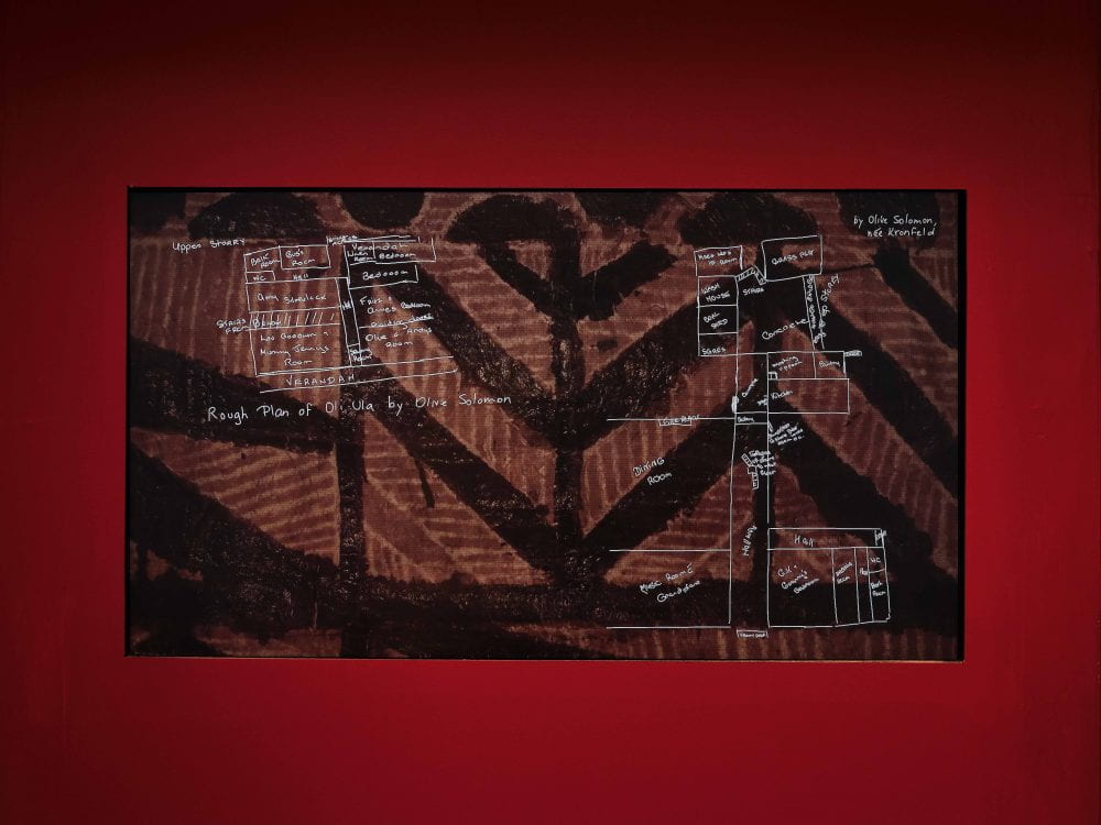A film screen embedded into a bright red wall. The film shows a floor map for the villa, against a background of pattern deep brown tapa cloth.