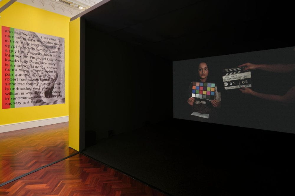 A film projected inside a large film booth. In the film a woman stares towards the camera, holding a colour chart against her face. A pair of hands from offscreen hold up a movie clapper.