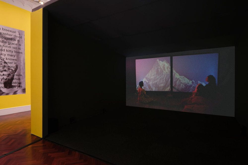 A film projected in a large film booth. In the film, two silhouetted figures sit on opposite ends of a two-screen projection, gazing up at an image of a snow covered mountain at sunset.