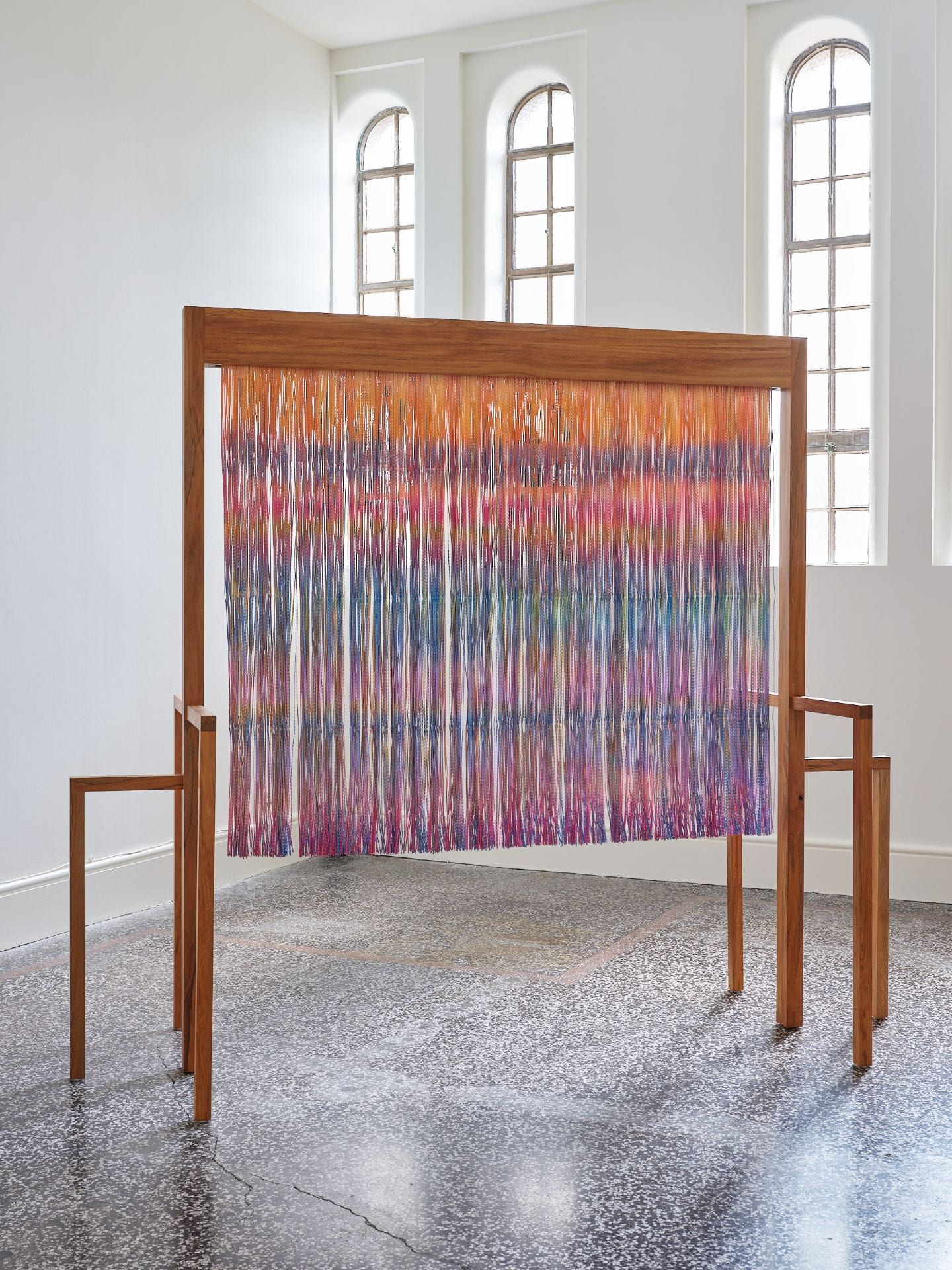 A rectangular wooden structure stands in the centre of a white gallery space. A veil of orange, blues, purples and pinks hangs from the structure. Windows at the back of the room let natural light in.